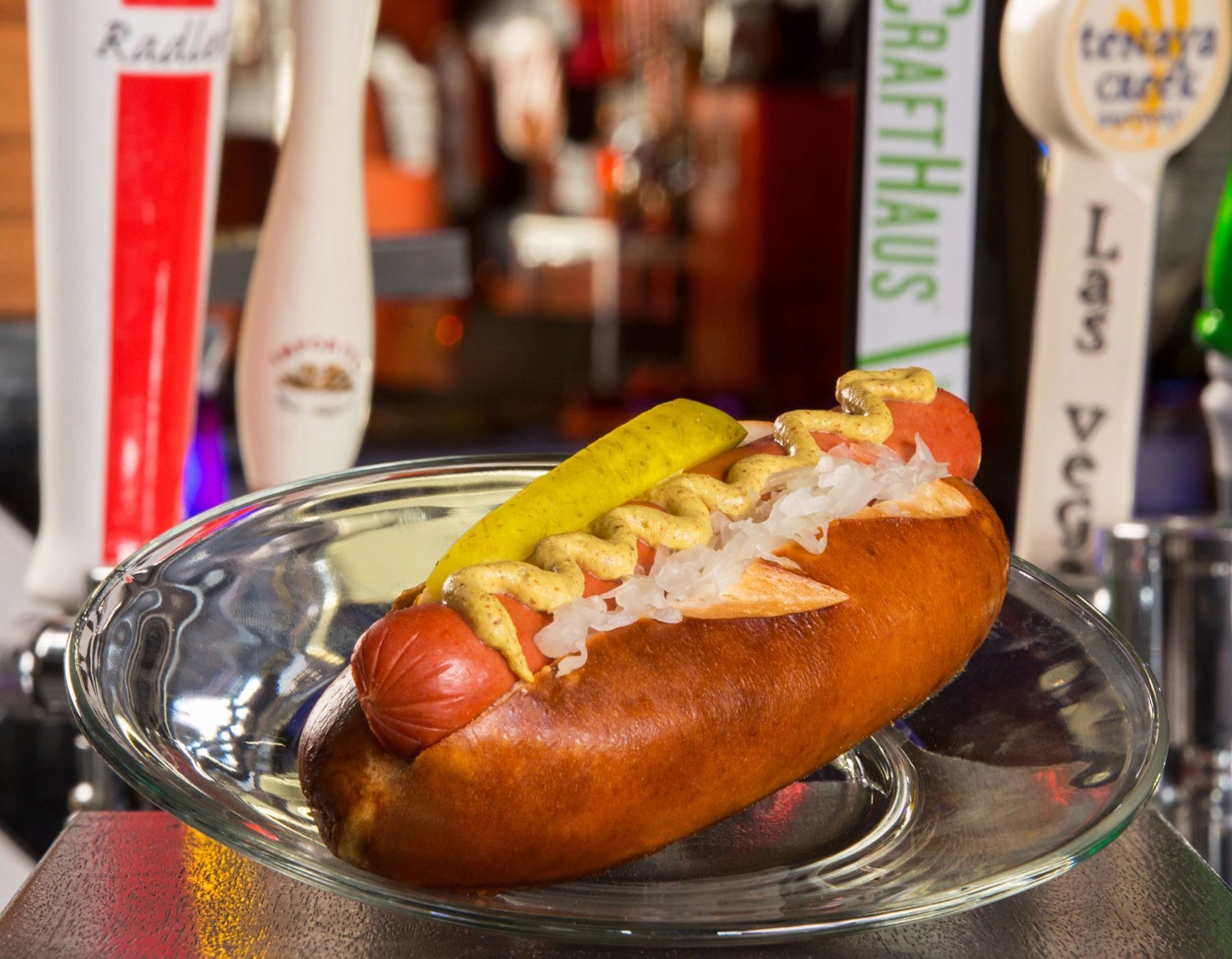 A grilled hot dog with sauerkraut and mustard on a glass plate in front of a row of craft beer taps