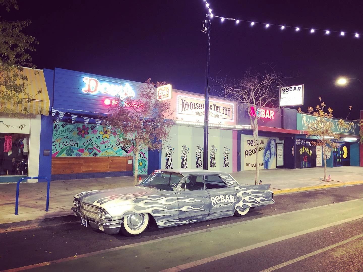 ReBAR's storefront row on the 18b strip at night, with the lowrider parked out front.